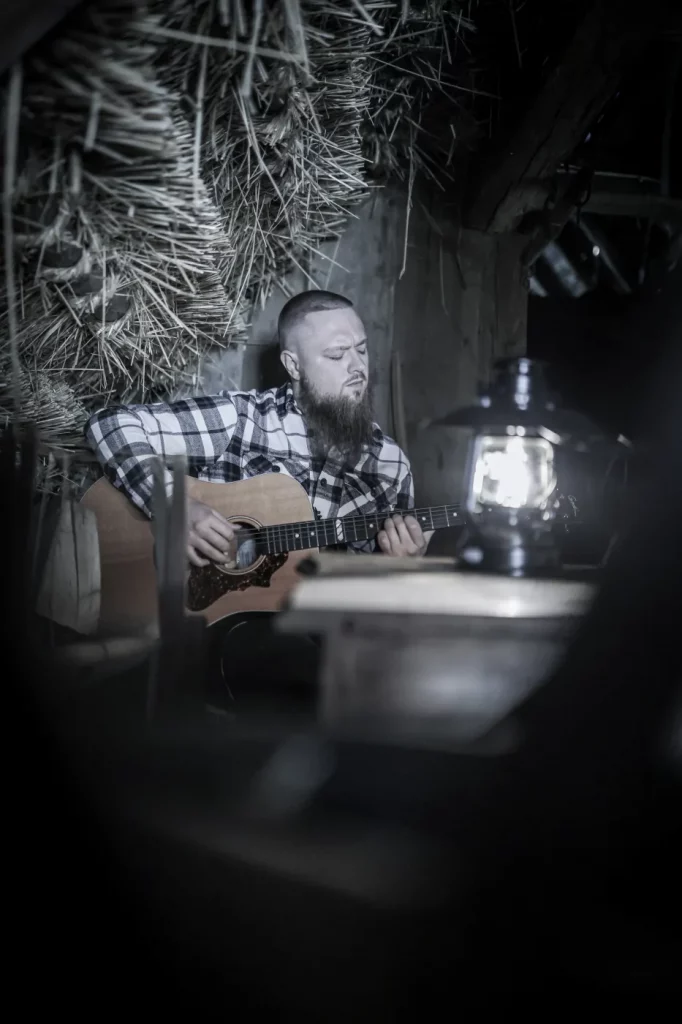 Nico Schwappacher (Zwischenlichten) mit Akustikgitarre. Schumriges Licht spendet eine kleine Lampe auf dem Tisch. 